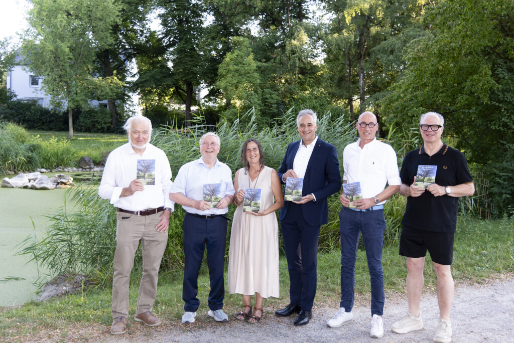 Sechs Beteiligte zeigen den 15. Band des Vitusbach vor einer begrünten Teichanlage.