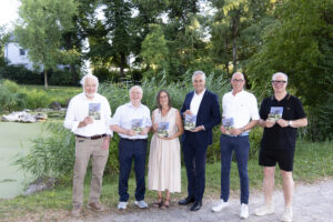 Sechs Beteiligte zeigen den 15. Band des Vitusbach vor einer begrünten Teichanlage.